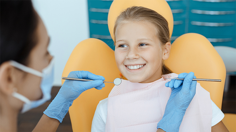 A child smiles during a dental checkup. She sits in a yellow chair. A staff member can be seen partially in the foreground.