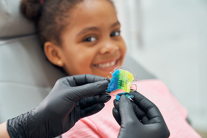 A metal retainer is shown; it is blue and yellow and held by two gloved hands. In the background, a child orthodontic patient smiles.