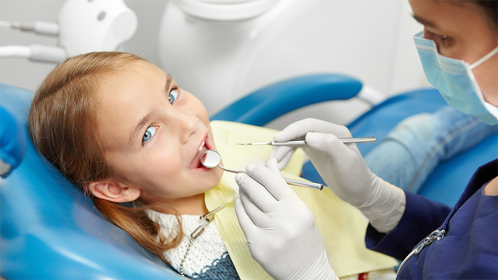 A child looks at the camera while a staff member examines their teeth.